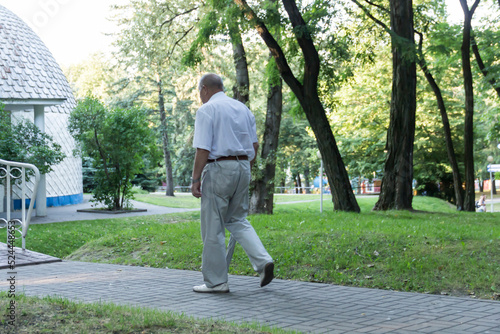 An old, stooped man walks sullenly and alone along the path in the park with a gray umbrella instead of a cane. photo