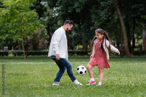 Positive asian kid and parent playing football on meadow in park.
