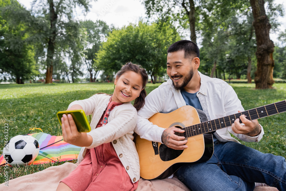 Cheerful asian girl taking selfie with dad playing acoustic guitar near flying kite and football in park.