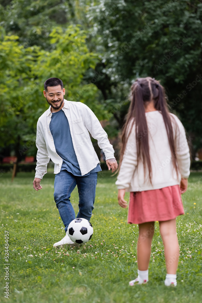 Cheerful asian dad playing football with blurred daughter in park. Cheerful asian dad playing football with blurred daughter in park.