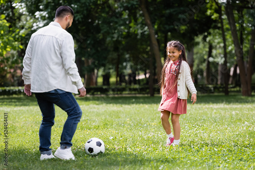 Smiling asian child playing football with father in summer park.