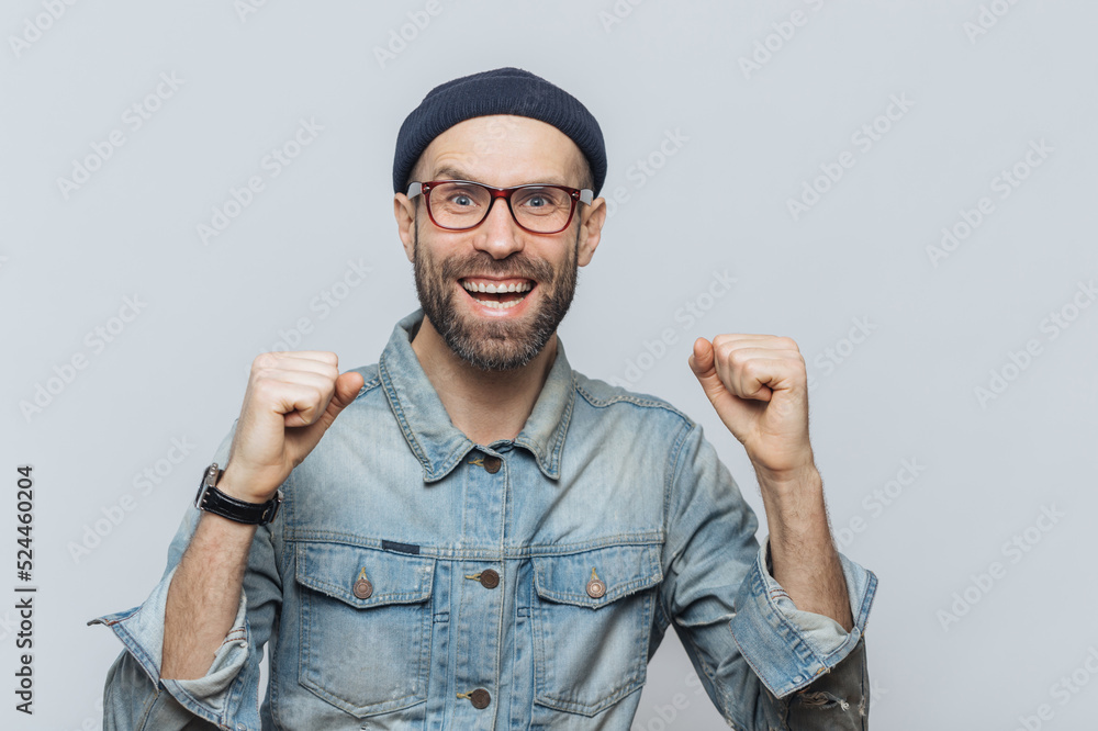 Overjoyed male rejoices his success, clenches fists, looks joyfully into camera, isolated over grey background. Bearded hipster guy raises hand with triumph. Middle aged man feels like champion Overjoyed male rejoices his success, clenches fists, looks joyfully into camera, isolated over grey background. Bearded hipster guy raises hand with triumph. Middle aged man feels like champion