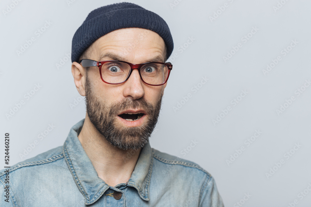 Suprised terrified unshaven male stares at camera and keeps mouth widely opened, wears fashionable clothing, expresses great shock as hears unexpected news from interlocutor, isolated on grey wall Suprised terrified unshaven male stares at camera and keeps mouth widely opened, wears fashionable clothing, expresses great shock as hears unexpected news from interlocutor, isolated on grey wall