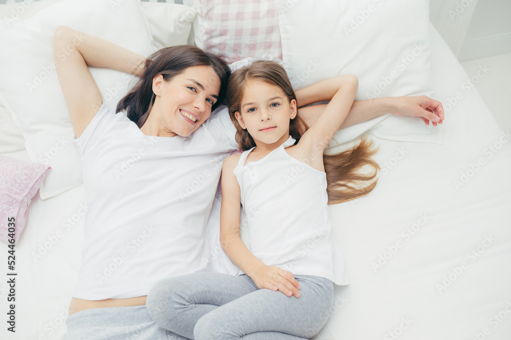 Top view of loving young mother with pleasant smile lies near her small daughter, going to have sleep, enjoy calm domestic atmosphere in bedroom, look directly at camera with joyful expressions Top view of loving young mother with pleasant smile lies near her small daughter, going to have sleep, enjoy calm domestic atmosphere in bedroom, look directly at camera with joyful expressions