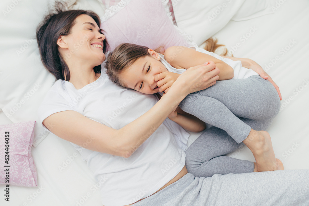 Delighted female child giggles joyfully as plays with her mother in comfortable bed, have positive smiles on faces, wear pyjamas, have good mood after awakening. People, leisure and sleeping concept Delighted female child giggles joyfully as plays with her mother in comfortable bed, have positive smiles on faces, wear pyjamas, have good mood after awakening. People, leisure and sleeping concept
