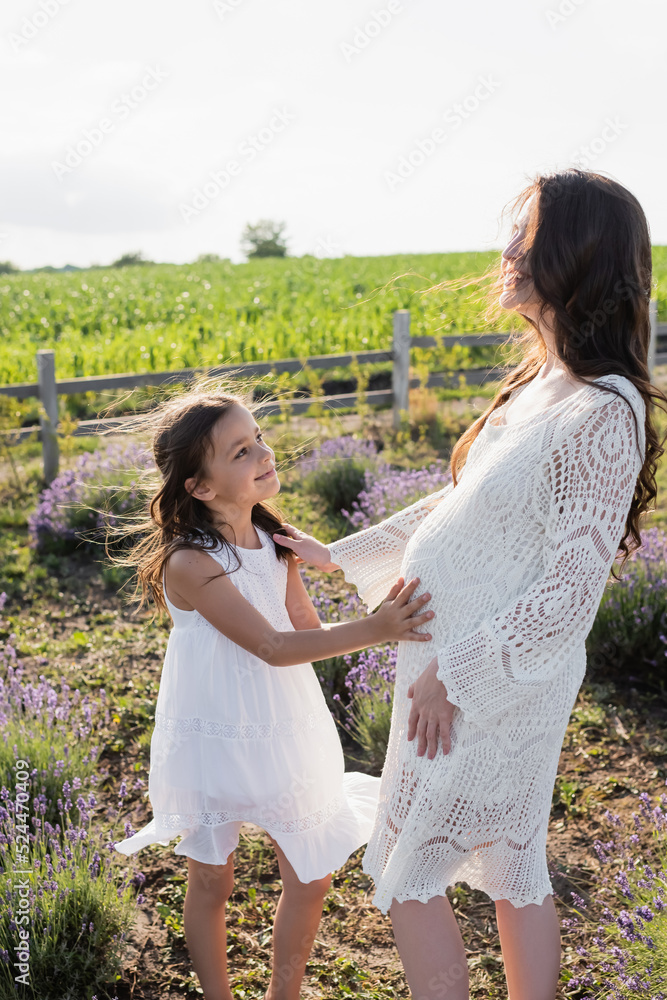 Naklejka premium smiling girl touching tummy of pregnant mom in field.
