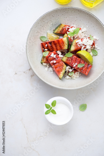 Plate with grilled watermelon batons, grated cheese and yogurt, above view on a light-beige marble background, vertical shot with space