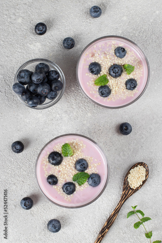 Thick yogurt with blueberries and sesame seeds in glasses, as well as in wooden spoon on gray background, Top view