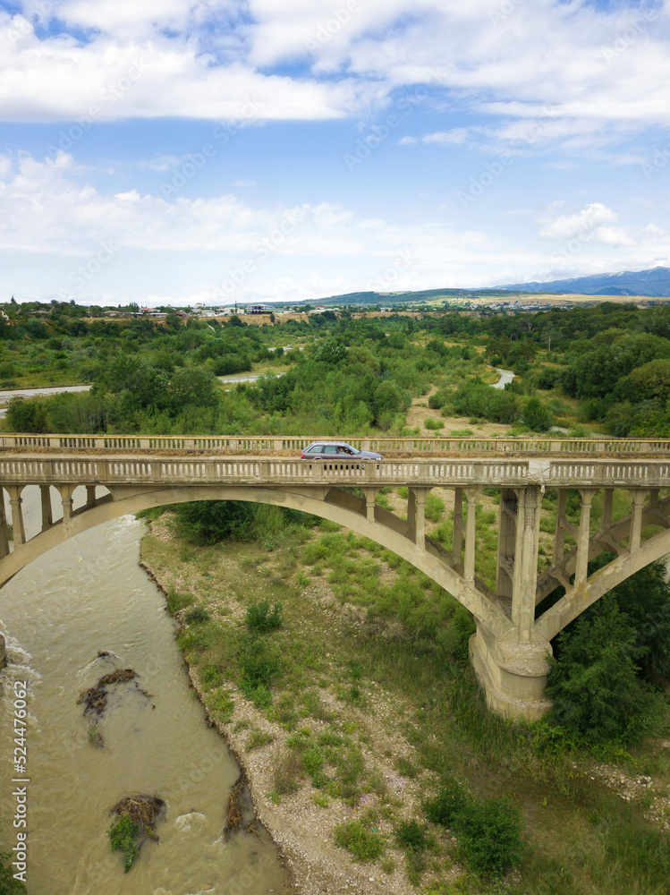 Car rides on an old arch bridge over a mountain river on a summer day, a side view from a drone