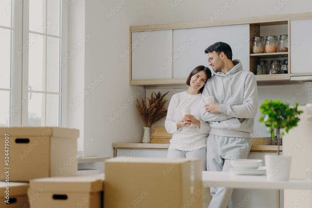 Photo of delighted relaxed husband and wife pose near modern kitchen furniture, have glad expressions, drink takeaway coffee, surrounded with cardboard boxes during relocation day. Mortgage. Photo of delighted relaxed husband and wife pose near modern kitchen furniture, have glad expressions, drink takeaway coffee, surrounded with cardboard boxes during relocation day. Mortgage.
