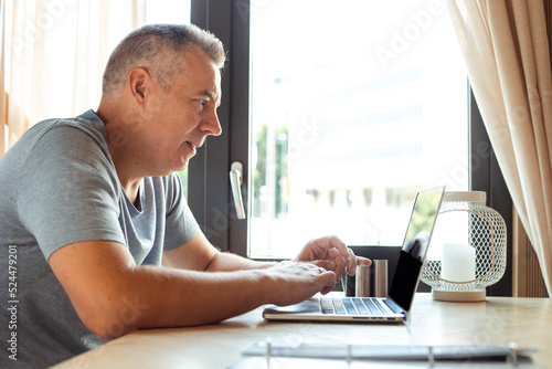 Elderly man sitting at table at home in summer, working on laptop, typing. Technology, working from home, freelance.