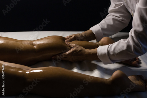 Uma terapeuta vestido de branco, fazendo massagem na perna de paciente deitado em maca, com o uso de óleo terapêutico.