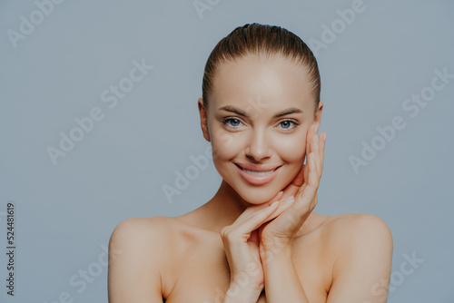Studio shot of pretty smiling woman touches face and enjoys flawless of skin  stands bare shoulders  has big forehead  poses against blue background  applies skin care cosmetics. Tenderness  makeup