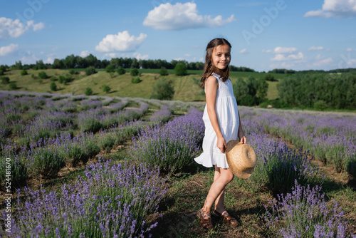 Fototapeta Naklejka Na Ścianę i Meble -  full length of smiling girl with straw hat standing in lavender field and looking at camera.