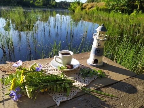 Still life with coffee breakfast, lighthouse toy, forest flowers on rustic wooden pier on riverside in sunny summer morning