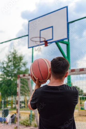 Rear view against the sky of a young guy in a black T-shirt who is outdoors on a sports field preparing to throw a basketball into a basket, copy space.