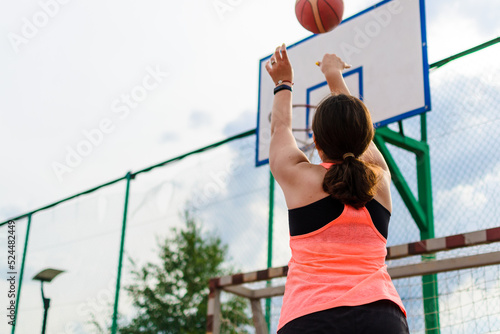 Woman in a bright tank top shooting a basketball at an outdoor court, with focus on determined posture and the basketball aimed towards the hoop, set against a clear sky.