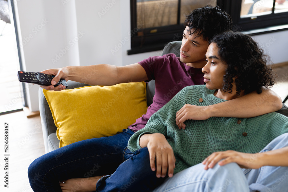Happy biracial lgbt male couple embracing on sofa in living room, watching tv Happy biracial lgbt male couple embracing on sofa in living room, watching tv