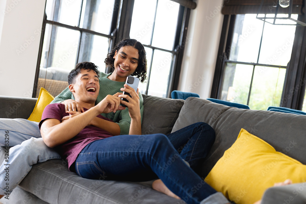 Happy biracial male lgbt couple sitting on sofa in living room, embracing and using smartphone Happy biracial male lgbt couple sitting on sofa in living room, embracing and using smartphone