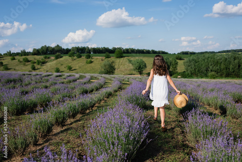 back view of brunette girl in white dress walking in lavender field with straw hat.