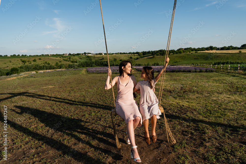 full length of woman and kid in summer dresses riding swing in meadow.