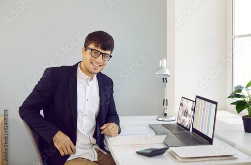 Portrait of a young business man or financial accountant at work. Happy handsome young man in a suit and eyeglasses sitting at his office desk with laptop computers, looking at the camera and smiling