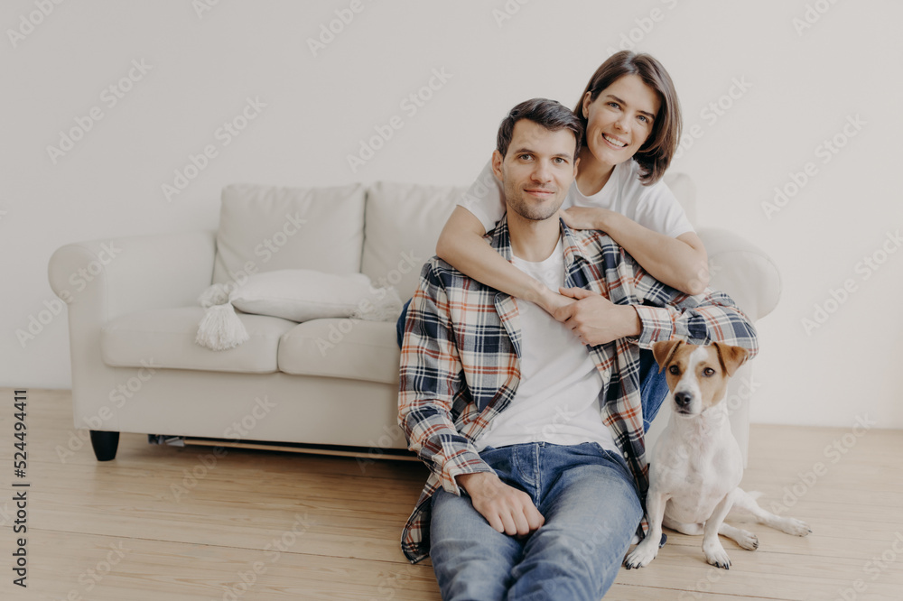 Photo of happy woman embraces her husband, poses on sofa, little dog sits on floor near owner, look all in camera with happy expressions, have romantic relationship. Family portrait. People and home Photo of happy woman embraces her husband, poses on sofa, little dog sits on floor near owner, look all in camera with happy expressions, have romantic relationship. Family portrait. People and home