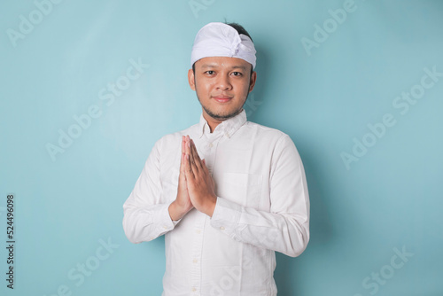 Smiling young Balinese man wearing udeng or traditional headband and white shirt gesturing greeting or namaste isolated over blue background