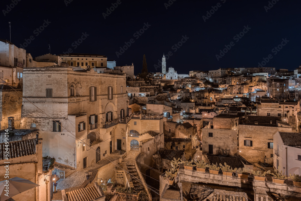 Fototapeta premium Matera by night, photos from the panoramic terrace. Photo from June 2022. You can see the basilica overlooking the town and the characteristic houses carved into the rock. Matera was the European capi