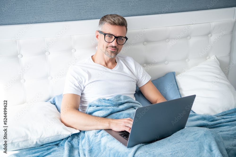 mature man virtual assistant in glasses working on laptop in bed mature man virtual assistant in glasses working on laptop in bed