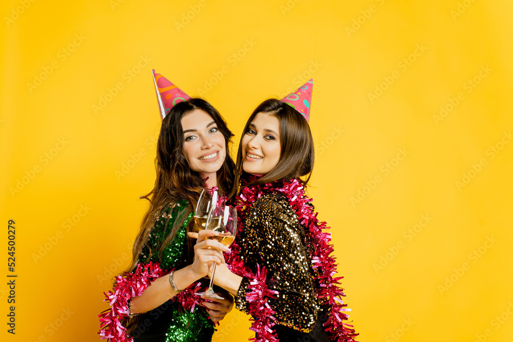 Young adult cheerful girls 20-25s in party hats and New Year's tinsel, hugging holding glasses of wine or champagne and looking at the camera on a yellow isolated background. Young adult cheerful girls 20-25s in party hats and New Year's tinsel, hugging holding glasses of wine or champagne and looking at the camera on a yellow isolated background.