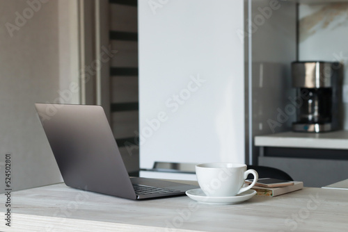 A laptop on a table with a cup, a notebook on the background, a refrigerator, a coffee maker