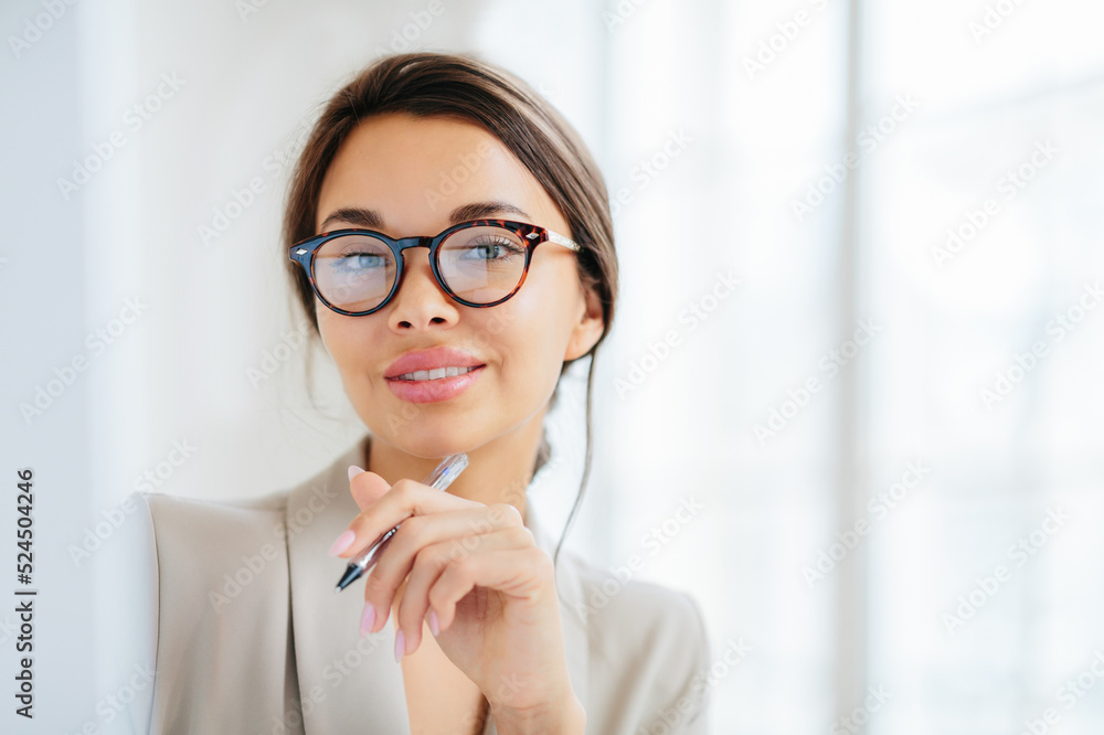 Headshor of attractive brunette woman holds pen, looks confidently at camera through spectacles, dressed in formal clothes, writes down notes, poses indoor. People, business and work concept Headshor of attractive brunette woman holds pen, looks confidently at camera through spectacles, dressed in formal clothes, writes down notes, poses indoor. People, business and work concept