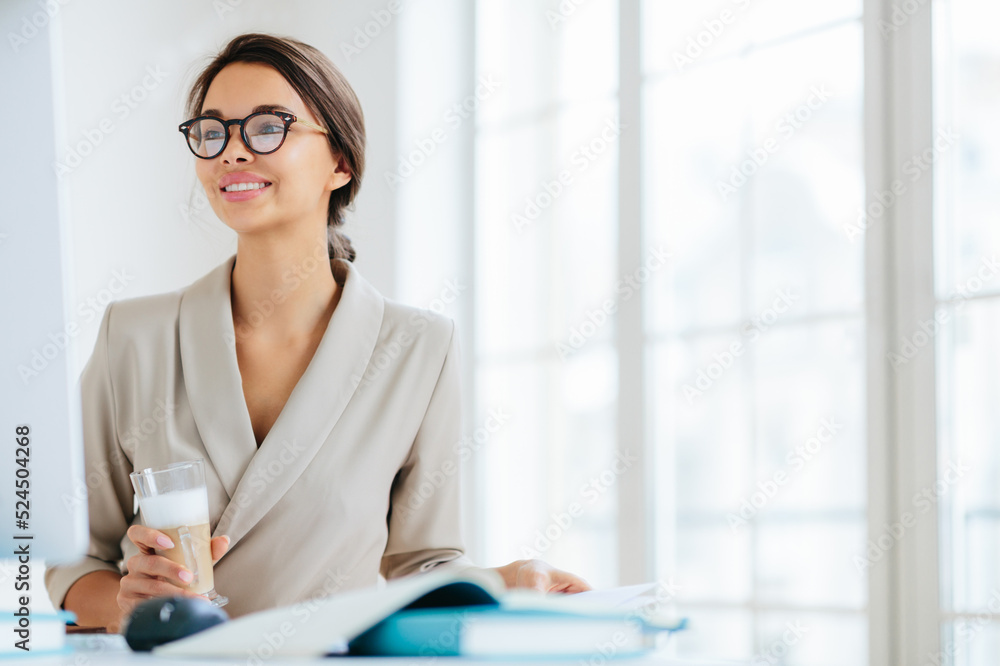 Positive woman works in office, sits at desktop, concentrated into monitor, drinks fresh milkshake, smiles and enjoys working, poses against office interior thinks about organization of business event Positive woman works in office, sits at desktop, concentrated into monitor, drinks fresh milkshake, smiles and enjoys working, poses against office interior thinks about organization of business event