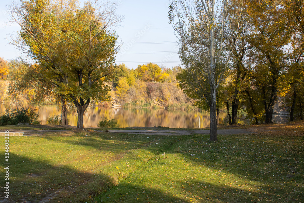 Autumn colors in sunny day on lake in city park. Landscape golden foliage and reflection of trees in calm water. Colorful plants on stone bank of pond. Autumn reflections. Fall bright. Beauty in natur