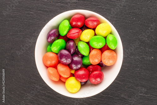 A lot of sweet colored candies in a white saucer on a slate stone, close-up, top view.