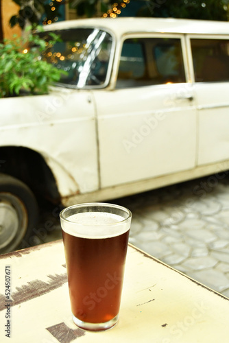 Cerveza Clara artesnal, servida en un vaso sobre una mesa, al fondo un automovil viejo y antiguo. photo