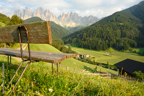 (Selective focus) Defocused park bench in the foreground with the Church of St. John (San Giovanni in Ranui) that stands out in the green meadows, in the heart of the beautiful Dolomitic landscape.. photo