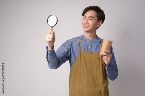 Portrait of young asian man wearing apron holding magnifying glass and paper cup over white background studio. © tonefotografia