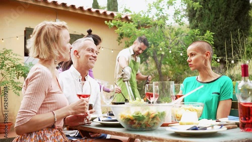 Three friends talking and having fun in a barbeque lunch. In the background a multiethnic couple cooking food. photo