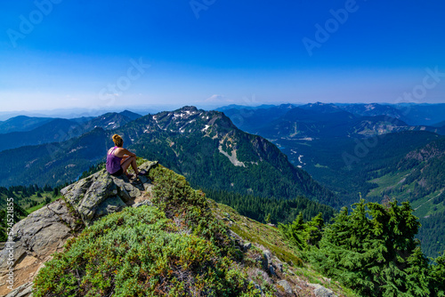 Adventurous athletic woman sitting on top of a mountain looking out at a mountain range on a beautiful sunny day in the Pacific Northwest. 