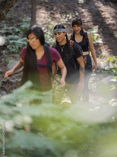 Vertical image of three Indigenous youth walking along a trail in a shaded forest 