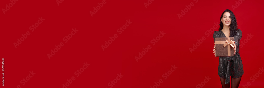 A pretty young girl in a black dress holds out a gift box standing on a red background. A pretty young girl in a black dress holds out a gift box standing on a red background.