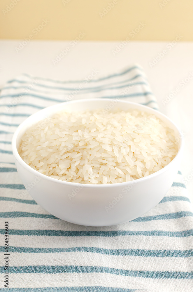 Dry camollino rice in a white ceramic bowl on a striped kitchen towel. Asian food concept. Healthy Eating. Copy space. Selective focus. Vertical orientation. Dry camollino rice in a white ceramic bowl on a striped kitchen towel. Asian food concept. Healthy Eating. Copy space. Selective focus. Vertical orientation.