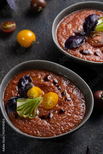 Traditional spanish cold tomato soup gazpacho in a bowl served with basil and cucumber on black textured table with fresh ingredients around. Top view with copy space. Dark mood healthy vegan dish.