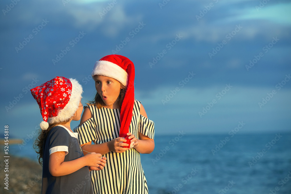 Obraz premium two sister girls in santa claus hats have fun on the seashore at sunset, one girl tells something emotionally to the other