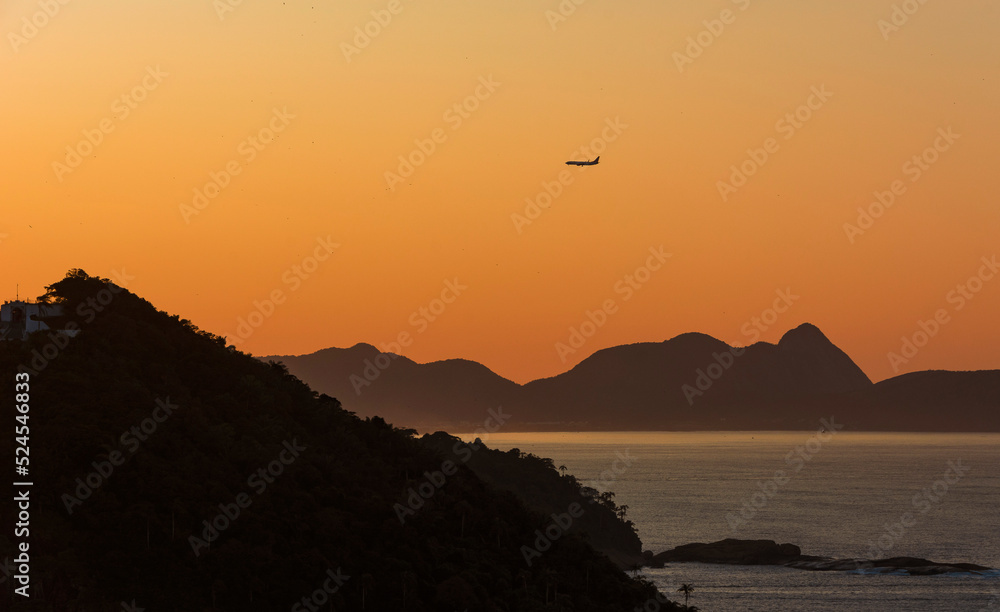 Obraz premium Airplane flying over the mountains at sunrise. Silhouettes and various shades of yellow and orange. Guanabara bay, Rio de Janeiro, Brazil. 