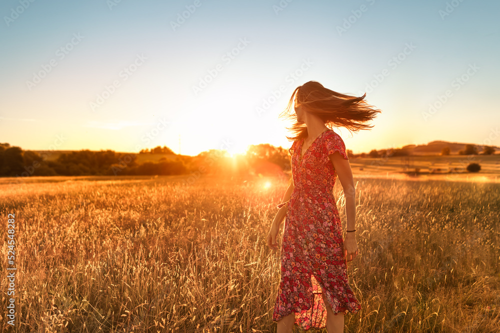 Naklejka premium Young woman walking in a field looking to the sunlight with wind blowing her hair. Beauty, freedom in nature concept. 