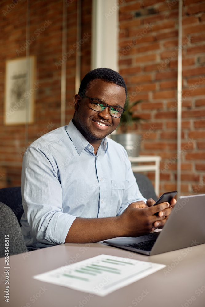 Happy black businessman texting on cell phone while working in office and looking at camera. Happy black businessman texting on cell phone while working in office and looking at camera.