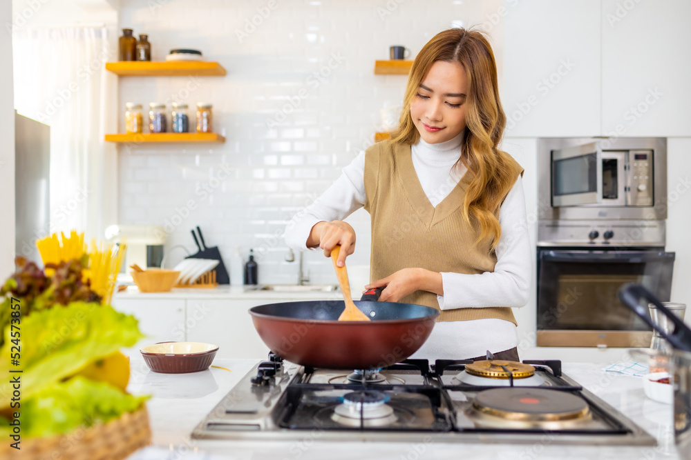 Young beautiful Asian woman enjoy cooking healthy food and pasta in cooking pan on stove in the kitchen at home. Happy female having dinner meeting party celebration with friends on holiday vacation.